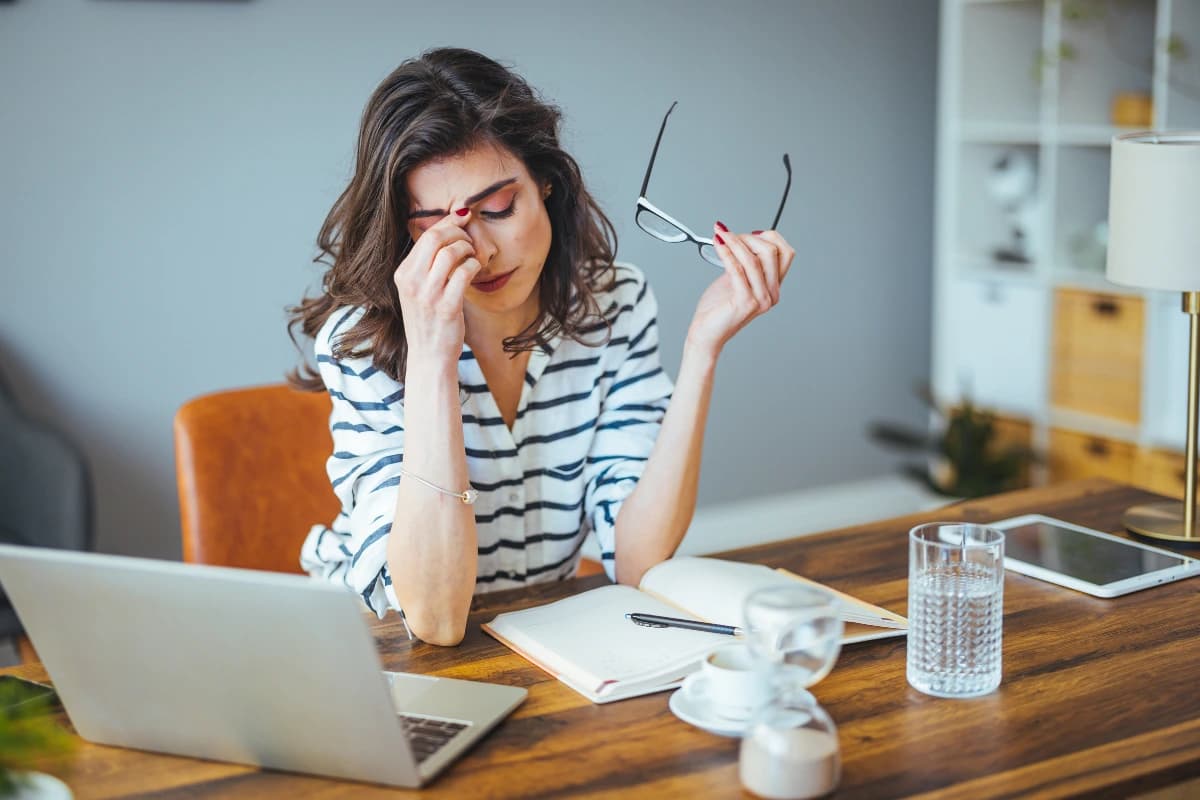 A tired woman holding glasses, pinching her nose in front of a laptop and notebook.