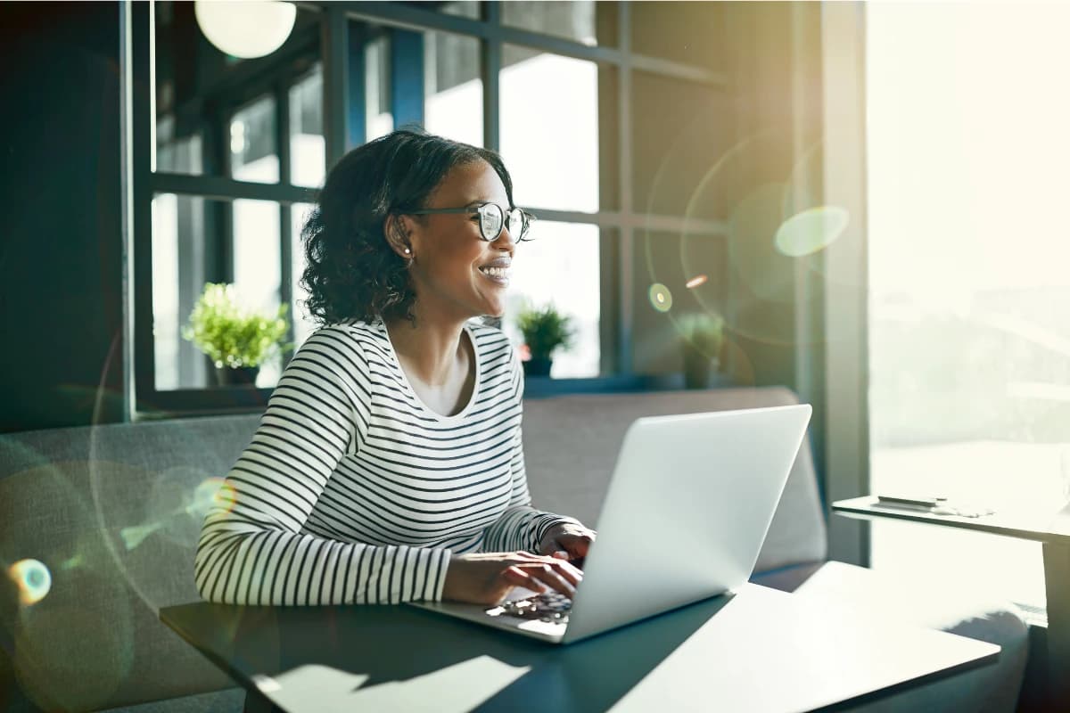 Person smiling while working on a laptop in a sunlit, modern workspace