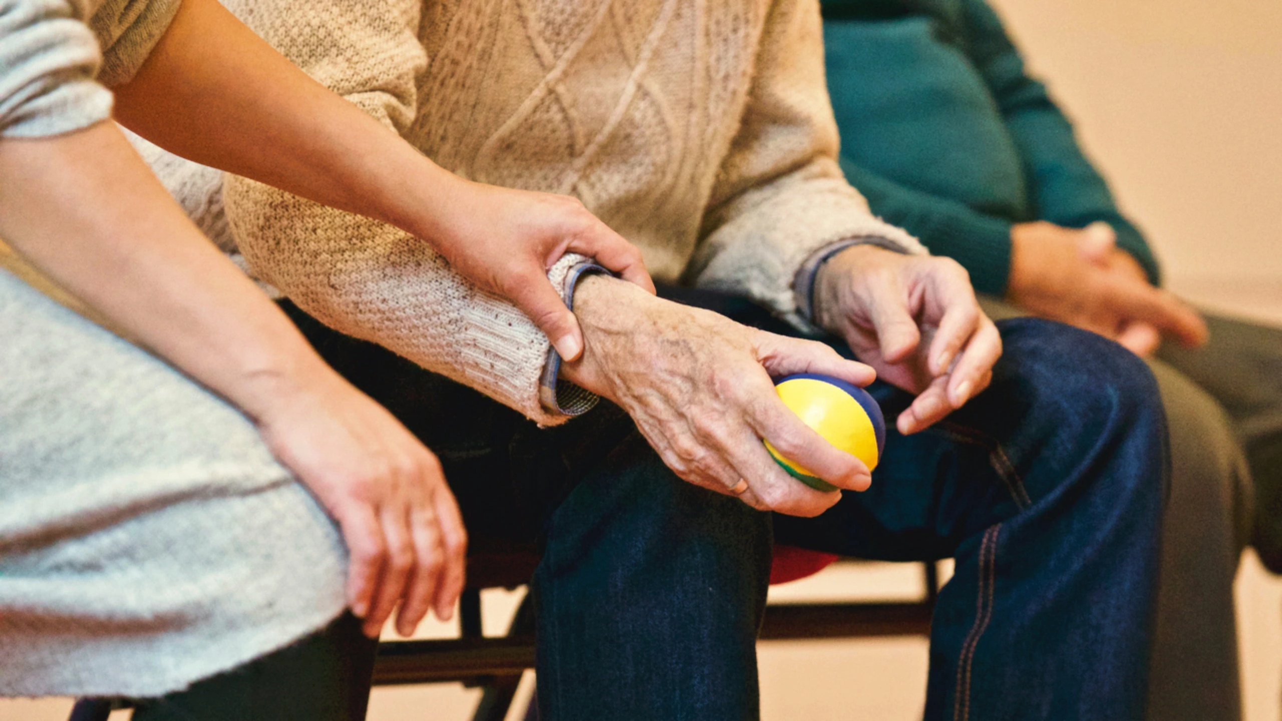 The image shows a caregiver gently supporting an elderly person holding a stress ball.